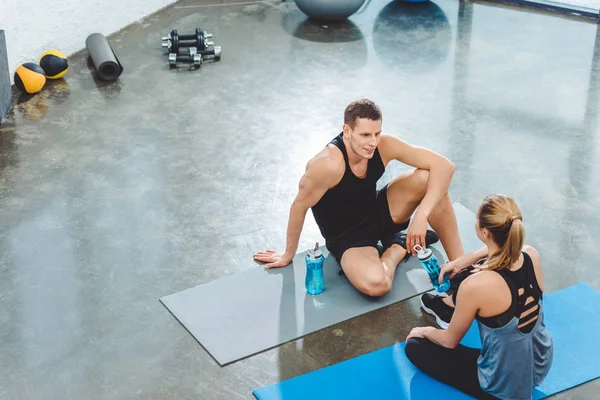 Vista de ángulo alto de deportista y deportista con botellas de agua sentados en colchonetas de yoga y mirándose en el gimnasio - foto de stock