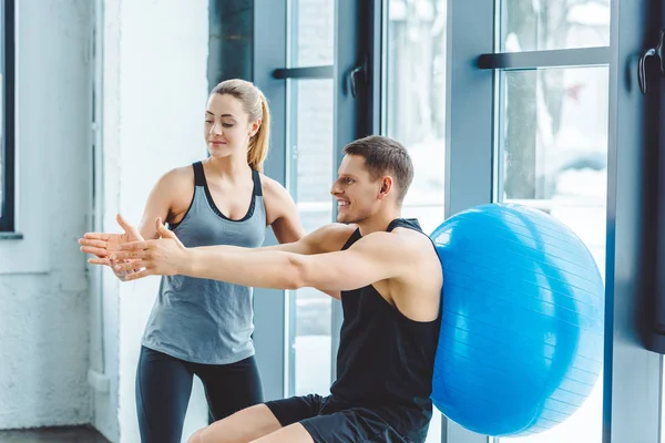 Donna che aiuta l'uomo sorridente che esercita con la palla di idoneità in palestra — Foto stock