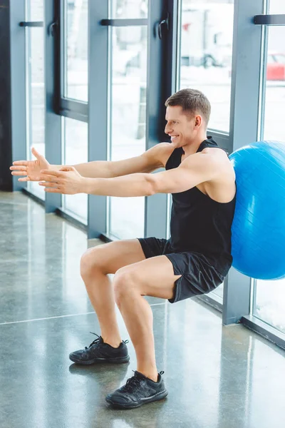 Vue latérale du sportif souriant faisant de l'exercice avec balle de fitness dans la salle de gym — Photo de stock