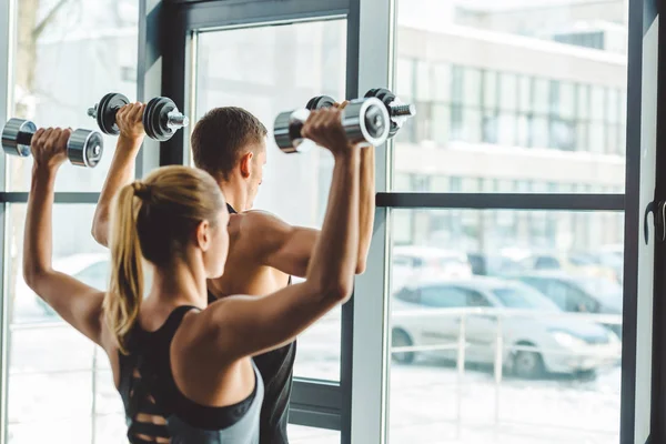 Rückansicht eines jungen Mannes und einer jungen Frau, die aus dem Fenster schauen, während sie im Fitnessstudio mit Kurzhanteln trainieren — Stockfoto