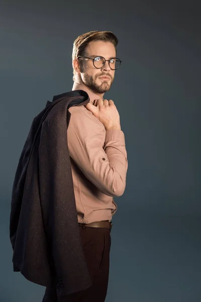 Портрет красивого стильного молодого чоловіка в окулярах, що тримає піджак і позує ізольовано на сірому — стокове фото