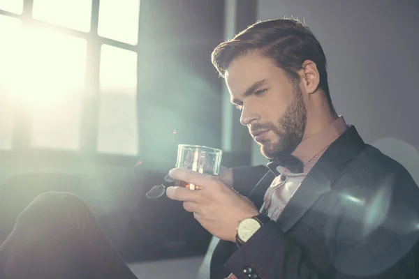 Handsome bearded young man holding eyeglasses and glass of whisky — Stock Photo
