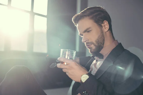 Beau jeune homme barbu tenant lunettes et verre de whisky — Photo de stock