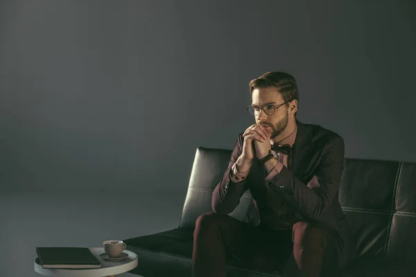 Pensivo jovem elegante homem em óculos sentado no sofá e olhando para longe — Fotografia de Stock