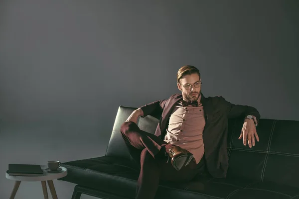 Handsome stylish young businessman in eyeglasses sitting on sofa and looking away on grey — Stock Photo