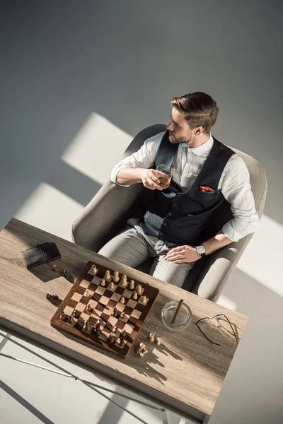 Высокий угол зрения молодого бизнесмена, смотрящего в сторону, когда он пьет виски и играет в шахматы — стоковое фото