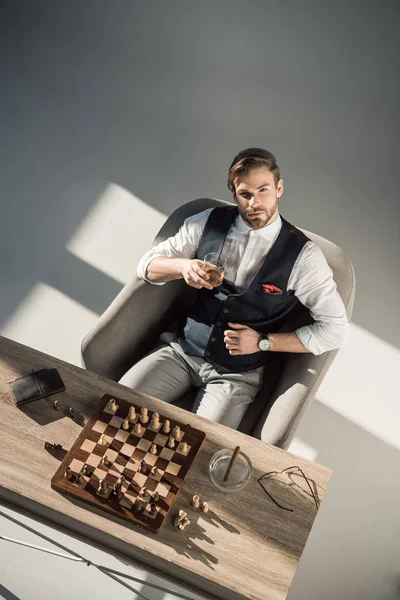 Aus der Vogelperspektive betrachtet blickt der junge Geschäftsmann in die Kamera, während er Whisky trinkt und Schach spielt — Stockfoto