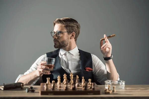 Joven con vaso de whisky fumando cigarro y mirando hacia otro lado mientras juega al ajedrez - foto de stock