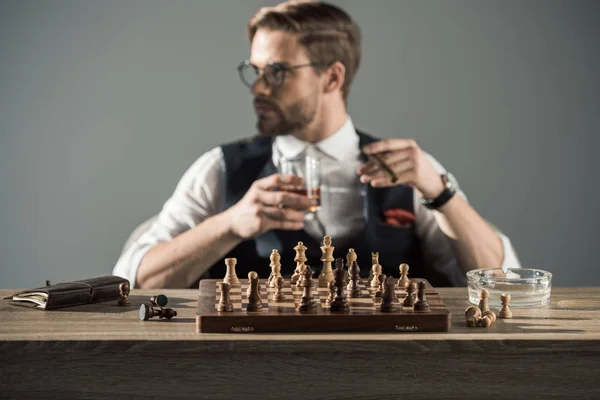 Крупный план шахматной доски с фигурами и человеком, пьющим виски и курящим сигару позади — стоковое фото