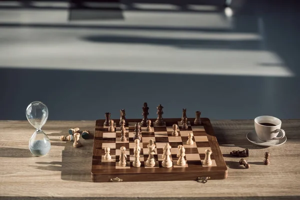 Close-up view of chess board with pieces, cup of coffee and sand clock on table — Stock Photo