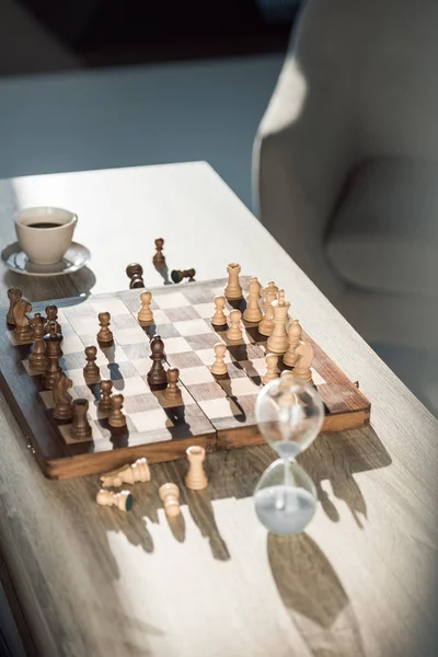 Enfoque selectivo de tablero de ajedrez con piezas, taza de café y reloj de arena en la mesa - foto de stock