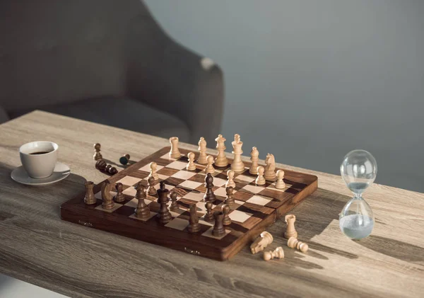 Vista de primer plano del tablero de ajedrez con piezas, taza de café y reloj de arena en la mesa - foto de stock