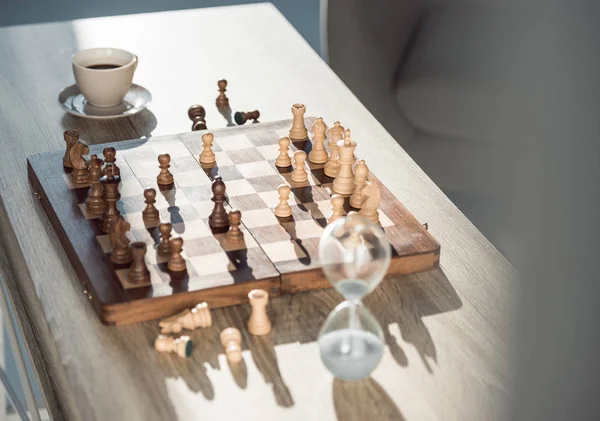 Foco seletivo de tabuleiro de xadrez com peças, xícara de café e relógio de areia na mesa — Fotografia de Stock