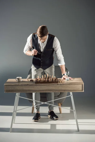 Jovem elegante segurando óculos e olhando para tabuleiro de xadrez com figuras — Fotografia de Stock