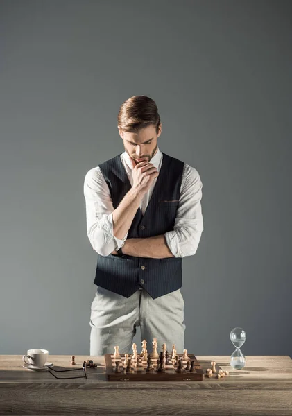 Pensativo joven mirando tablero de ajedrez con figuras y reloj de arena — Stock Photo