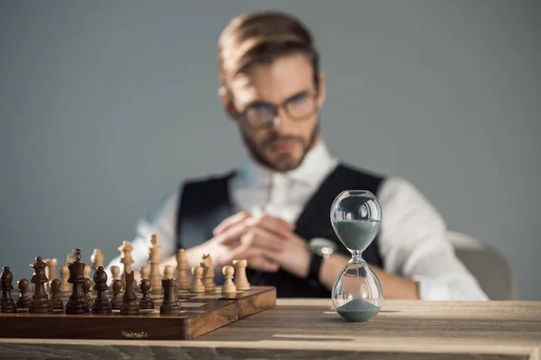 Vue rapprochée de l'échiquier avec des pièces et horloge de sable sur la table et homme d'affaires assis derrière — Photo de stock