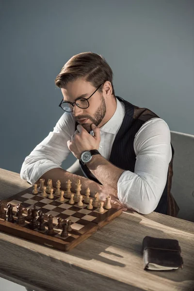 Stylish young businessman with hand on chin looking at chess board — Stock Photo