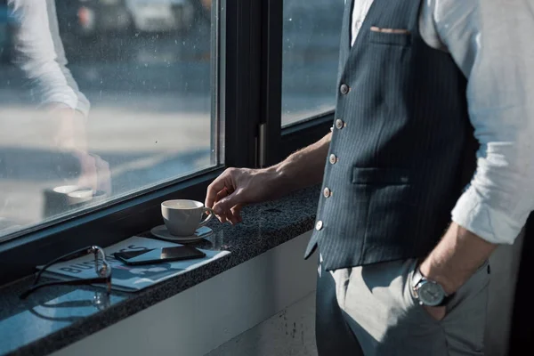 Sección media de hombre de negocios con la mano en el bolsillo beber café cerca de la ventana - foto de stock