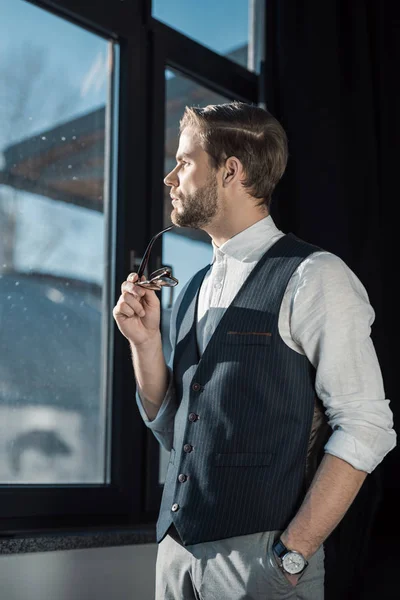Портрет задумчивого молодого бизнесмена, держащего очки и смотрящего в окно — стоковое фото