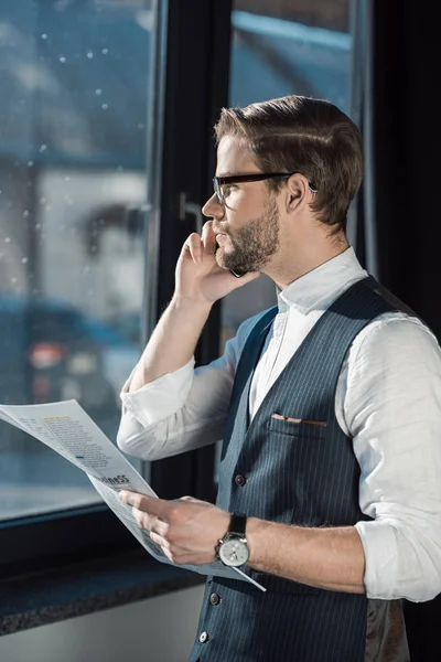 Portrait of stylish young businessman holding newspaper and talking by smartphone — Stock Photo