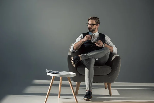 Schöner stylischer junger Mann sitzt im Sessel und schaut weg, während er Kaffee trinkt — Stockfoto