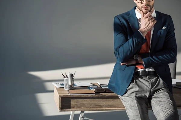 Обрезанный снимок стильного молодого бизнесмена с рукой в подбородке, сидящего на столе — стоковое фото