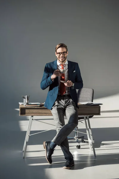 Lächelnder stilvoller Geschäftsmann mit einer Tasse Kaffee auf dem Tisch sitzend und in die Kamera blickend auf grau — Stockfoto