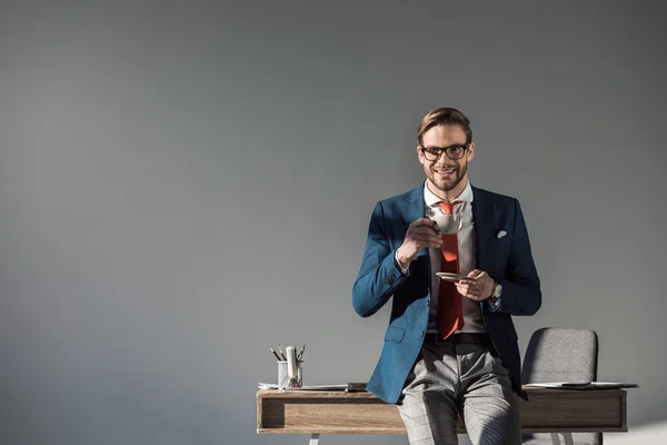 Улыбающийся бизнесмен с чашкой кофе сидит на столе и смотрит в камеру на сером — Stock Photo