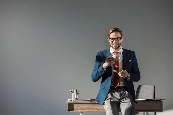 Smiling businessman with cup of coffee sitting on table and looking at camera on grey — Stock Photo