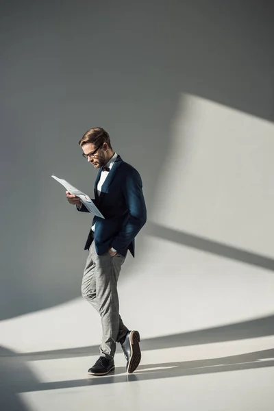 Vue pleine longueur de bel homme d'affaires élégant dans les lunettes de lecture journal sur gris — Photo de stock