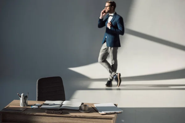 Selective focus of table with office supplies and open notebook and businessman talking by phone behind — Stock Photo