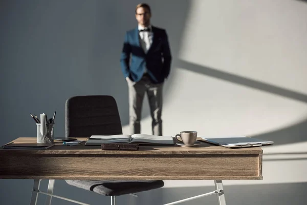Messa a fuoco selettiva del tavolo con forniture per ufficio e notebook aperto e uomo d'affari in piedi dietro — Foto stock