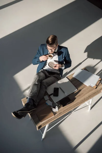 Vue grand angle de jeune homme d'affaires élégant dans les lunettes de vue boire du café sur le lieu de travail — Photo de stock