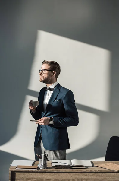 Stylish young businessman in eyeglasses drinking coffee at workplace — Stock Photo