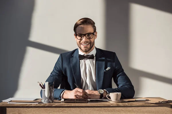 Bello giovane uomo d'affari sorridente alla macchina fotografica mentre seduto sul posto di lavoro — Foto stock