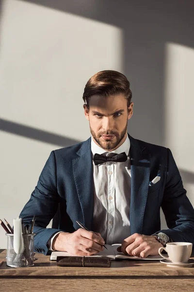 Schöner stylischer Geschäftsmann blickt in die Kamera, während er in Notizblock schreibt — Stockfoto