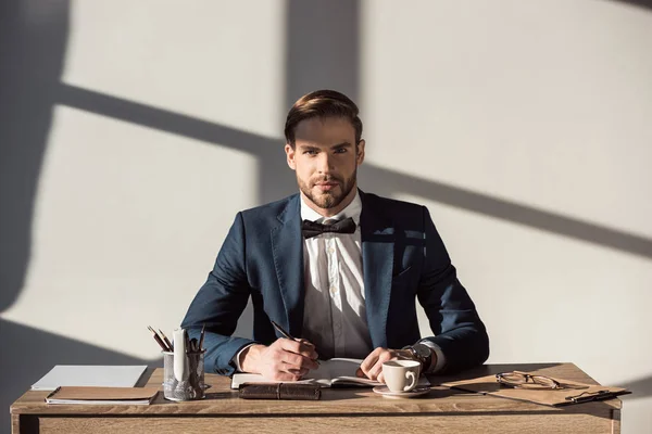 Schöner stylischer Geschäftsmann blickt in die Kamera, während er am Arbeitsplatz Tagebuch schreibt — Stockfoto