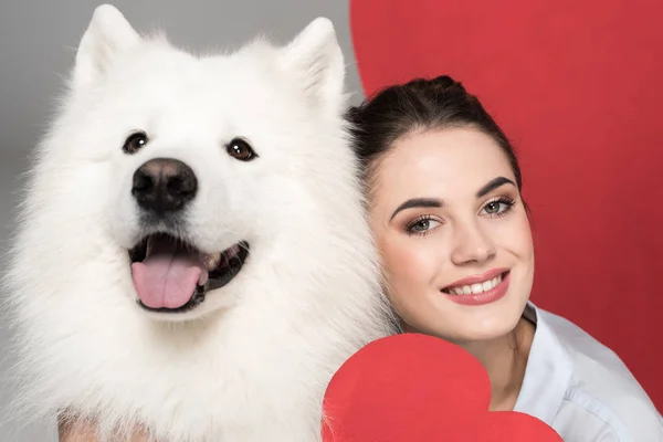 Fille souriante avec chien blanc et coeur en papier sur blanc, concept de Saint-Valentin — Photo de stock