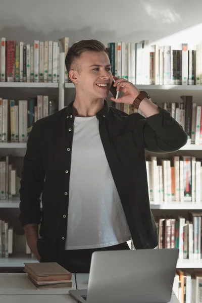 Estudiante guapo hablando por teléfono inteligente en la biblioteca - foto de stock