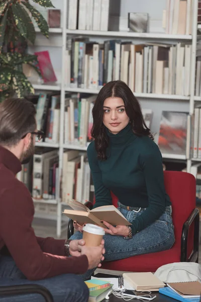 Hermosa chica sentada con libro en la biblioteca y mirando hacia otro lado - foto de stock