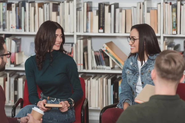 Улыбающиеся девушки сидят в библиотеке и смотрят друг на друга — стоковое фото