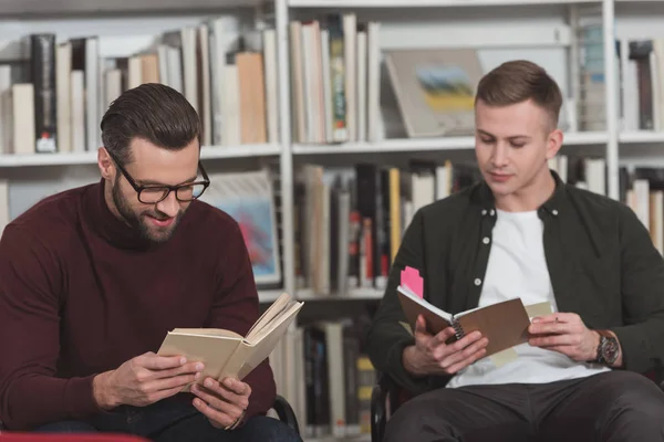 Hombres guapos sentados con libros en la biblioteca - foto de stock