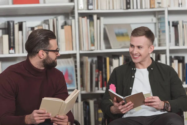 Homens bonitos sorridentes sentados com livros na biblioteca e olhando um para o outro — Fotografia de Stock