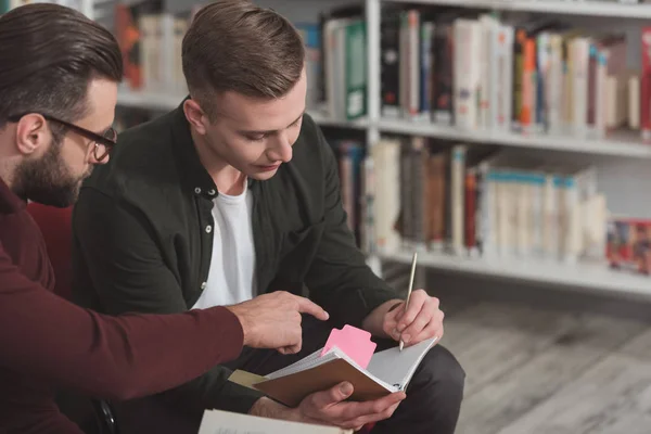 Mann zeigt in Bibliothek auf etwas im Notizbuch von Freunden — Stockfoto