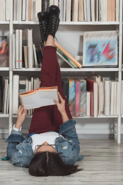 Menina afro-americana deitada no chão na biblioteca com pernas para cima e livro de leitura — Fotografia de Stock