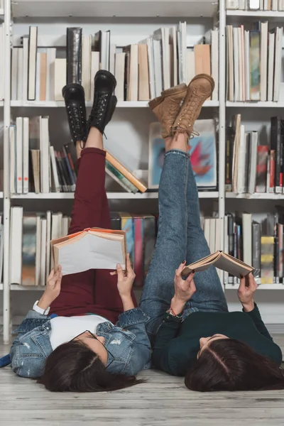 Amigos multiculturales tumbados en el suelo en la biblioteca con las piernas arriba y la lectura de libros - foto de stock