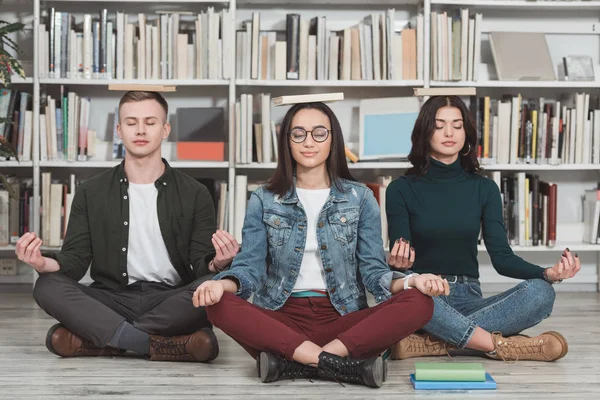Estudiantes multiculturales meditando con libros sobre cabezas en la biblioteca - foto de stock