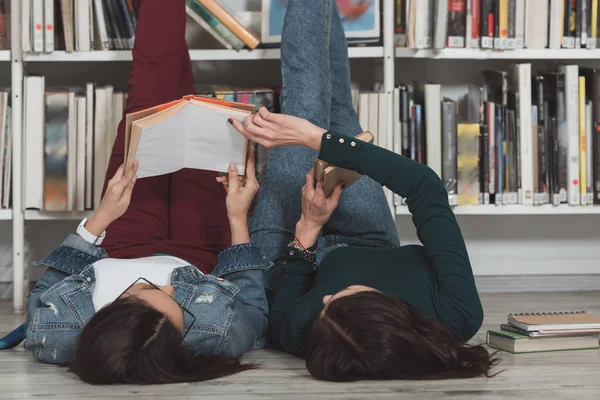 Amis multiculturels couchés sur le sol dans la bibliothèque avec les jambes en l'air et lecture livre — Photo de stock