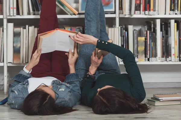 Multicultural friends lying on floor in library with legs up and reading book — Stock Photo