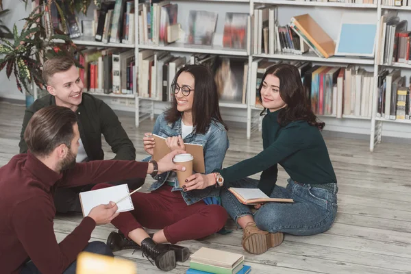 Amigos multiculturales felices sentados en el piso en la biblioteca y compartiendo café - foto de stock